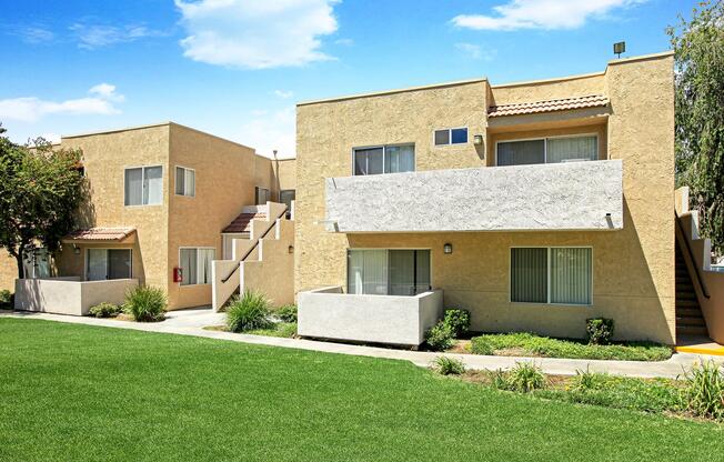 Two modern beige apartment buildings with balconies are situated in a landscaped area featuring green grass. The buildings have large windows and small staircases leading to the entrances. Bright blue sky with scattered clouds can be seen overhead, creating a sunny, inviting atmosphere.