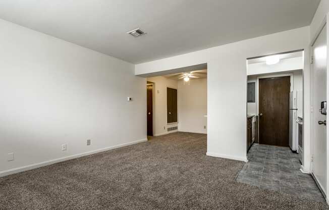 an empty living room with white walls and carpeting and a door to a kitchen