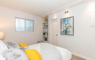 A cozy bedroom featuring a light-colored bed with yellow throw pillows and a soft gray blanket. There's a modern piece of abstract art on the wall and a shelf with decorative items. A small window allows natural light, and the walls are painted in neutral tones, creating a calm and inviting atmosphere.