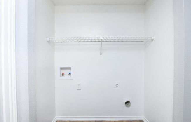 A minimalist empty closet featuring a white wall, a wire shelf mounted on the wall, and a small utility panel with pipes. The floor is wooden, and there is a vent or hole near the baseboard. The overall space appears clean and well-lit.