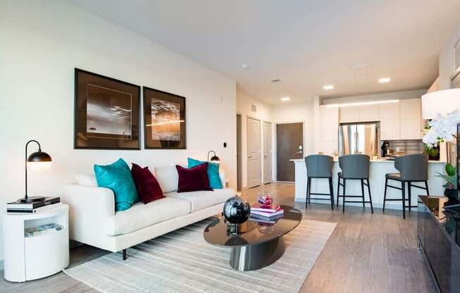 A modern living room with a white sofa, a coffee table, and a dining area in the background.