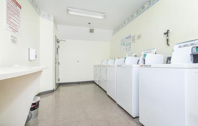 A clean and well-lit laundry room featuring several white washing machines aligned against the wall. There is a counter space on the left side and a door at the far end. Fresh, neutral-colored walls and a tiled floor contribute to the bright and tidy atmosphere.