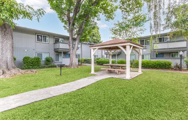 A well-maintained courtyard featuring a gazebo with a picnic table on a grassy area, surrounded by trees and apartment buildings. The scene is bright and inviting, perfect for outdoor gatherings or relaxation.
