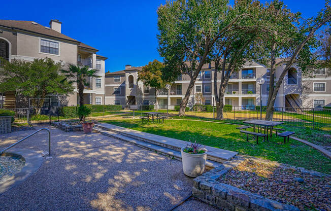 A spacious outdoor picnic area at Saxony at Chase Oaks Apartments in Dallas, TX, featuring picnic tables, a grassy lawn, and trees, offering a peaceful space for residents to gather.