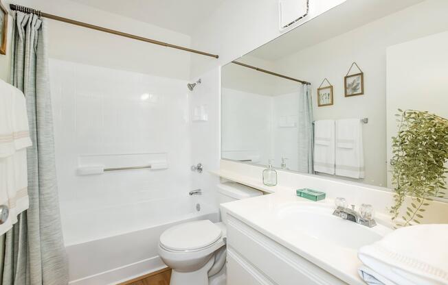 A modern bathroom featuring a bathtub with a shower, a large mirror, and a vanity with a sink. The decor includes light-colored walls, a shower curtain, and plants. Towels are neatly hung, and there are small decorative frames on the wall, creating a clean and inviting space.
