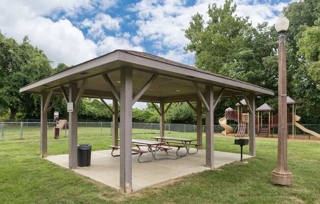 A covered picnic area with tables and a trash can is surrounded by a playground and trees.