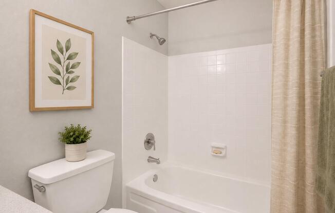 A clean and modern bathroom featuring a white bathtub with a shower, a light-colored shower curtain, a wall-mounted showerhead, a potted plant on the countertop, and a framed botanical print on the wall. The overall design is minimalistic and airy, with soft neutral tones.