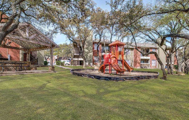A playground with a red swing set is surrounded by trees and a building in the background.