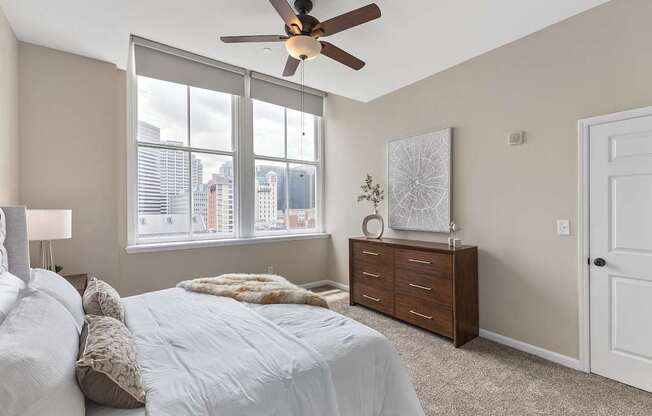 A bedroom with a bed, a ceiling fan, and a window overlooking a cityscape.