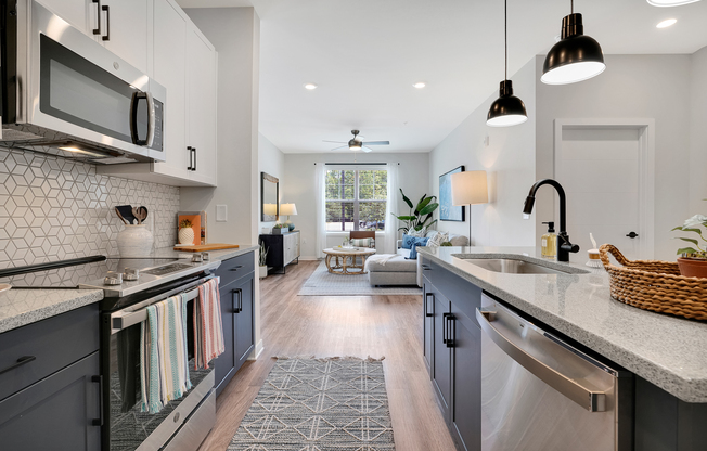 Modern kitchen with gray cabinetry, stainless steel appliances, and a geometric backsplash. A dining area with a rug leads to a bright living room featuring a sofa, coffee table, and large windows. Decorative items include a plant and a wicker basket on the counter. Bright lighting enhances the airy atmosphere.