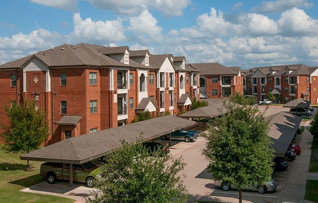 A community parking area here at Landing at Mansfield with brick apartment buildings, covered carports, parked cars, and green trees lining the drive under a bright, partly cloudy sky.