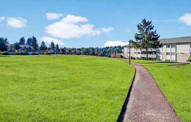A peaceful outdoor walking path here at Andorra winding through expansive green lawns and manicured landscaping, with a smooth paved walkway leading past mature trees, open grassy areas, and nearby apartment buildings, creating a scenic setting for daily walks, fresh air, and enjoying the surrounding community views.
