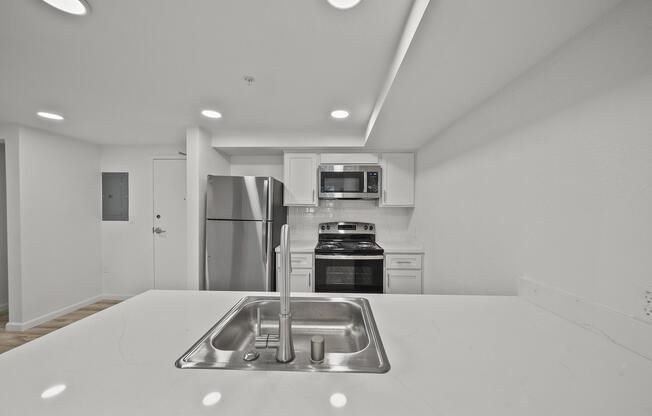 Modern kitchen with a central island featuring a sink. Stainless steel appliances, including a refrigerator and an oven, are visible in white cabinetry. The space is well-lit with recessed ceiling lights and has a clean, minimalist aesthetic, highlighted by light-colored walls and wooden flooring.