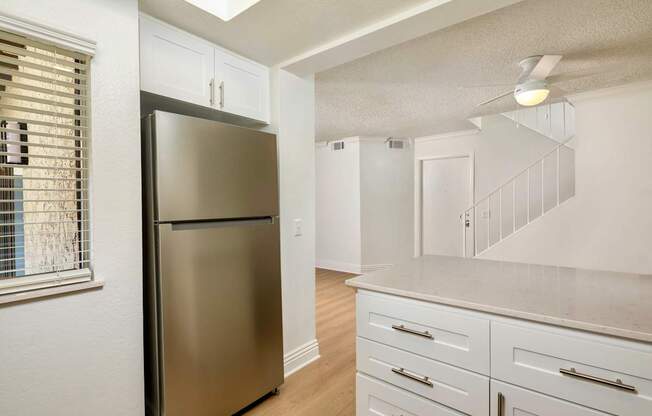 A stainless steel refrigerator in a kitchen with white cabinets and a window with blinds.