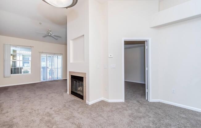 Spacious interior of a living room with light beige walls and carpet. Features include a corner fireplace, ceiling fan, and large windows allowing natural light. An open doorway leads to another room, while sliding doors provide access to a balcony or patio.