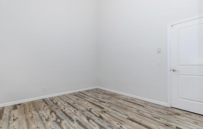 Empty room with light gray walls and a wooden floor. The space features a single white door on the right side, and there are no furnishings or decorations present, creating a minimalist and spacious appearance. Natural light may be entering, contributing to the bright atmosphere.