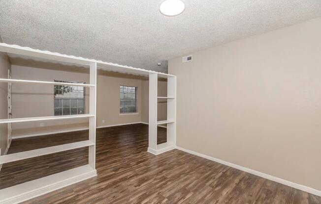 A spacious, empty room featuring light brown walls, laminate flooring, and a ceiling light. There are built-in white shelves separating areas, and two windows with views of the outside. The room is designed for versatile use, providing ample natural light.