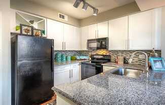 A kitchen here at Lakewood on the Trail with white cabinets, granite countertops, black refrigerator and range, mosaic tile backsplash, and teal decor accents, creating a bright, modern, and inviting cooking space.