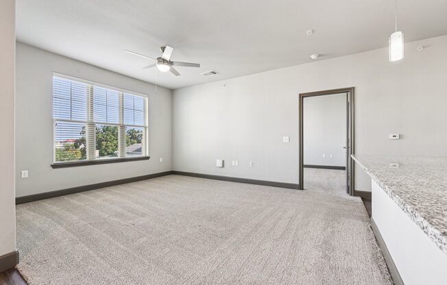 A spacious room with a carpeted floor, a ceiling fan, and a large window with blinds.