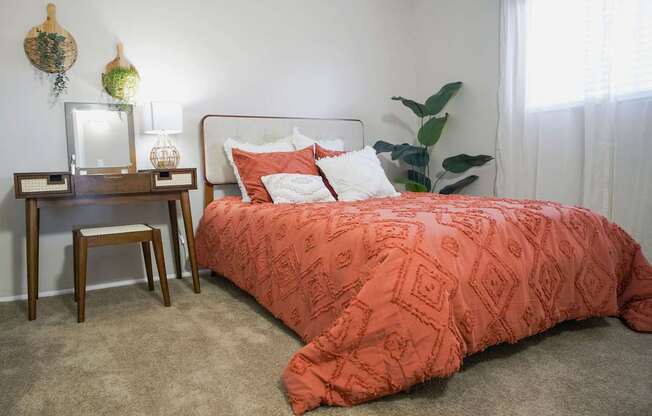 A bed with a red comforter and a white headboard sits in a room with a wooden nightstand and a plant.