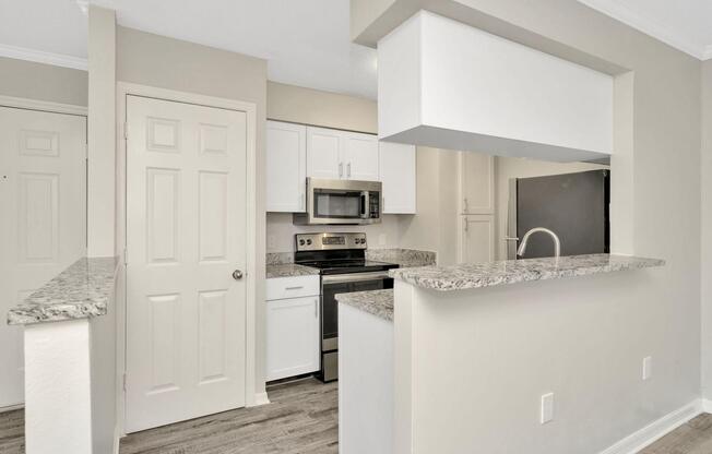 A modern kitchen featuring white cabinets, granite countertops, and stainless steel appliances. There is a small breakfast bar with a view into the kitchen, which includes a microwave, stove, and refrigerator. The walls are painted a light color, contributing to a bright and clean appearance.