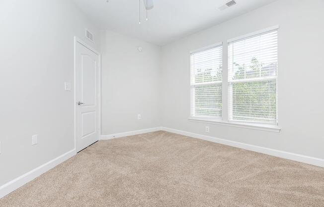 A bright, empty room with light gray walls and beige carpet. It features two large windows with white blinds, allowing natural light to flood in. A white door is visible on the left side, and the ceiling has a ceiling fan. The overall space appears clean and well-maintained, suitable for various uses.
