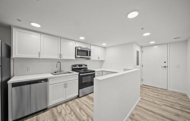 Modern kitchen featuring white cabinetry, stainless steel appliances including a refrigerator, stove, and dishwasher. The countertop is light-colored with a clean, minimalist design. The flooring is wood-like laminate, and overhead lights provide ample illumination. A door leads to the entryway.