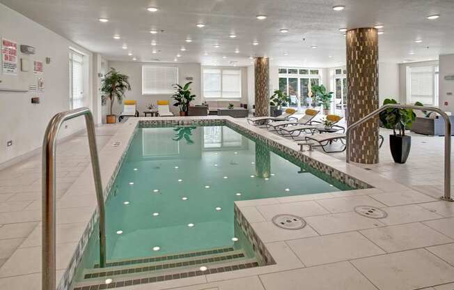 A bright indoor pool here at Heritage Hills featuring clear turquoise water, tiled pool surround, and stainless handrails for easy entry. The space includes cushioned lounge chairs with accent pillows, potted greenery, patterned columns, and large windows that allow natural light, creating a relaxing, resort-style atmosphere for year-round enjoyment.