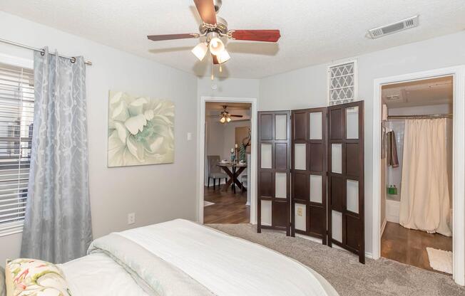 A cozy bedroom featuring a light-colored bed with a decorative floral pillow, a ceiling fan with wooden blades, and soft gray walls. To the left, there's a large floral painting and a room divider. A doorway leads to another room with a dining setup visible in the background. A bathroom area is also indicated.