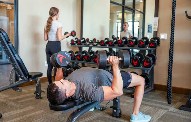 A man is working out in a gym with a woman in the background.