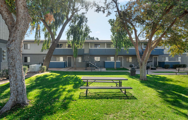 A picnic table is in the foreground of a grassy area with trees and apartment buildings in the background.