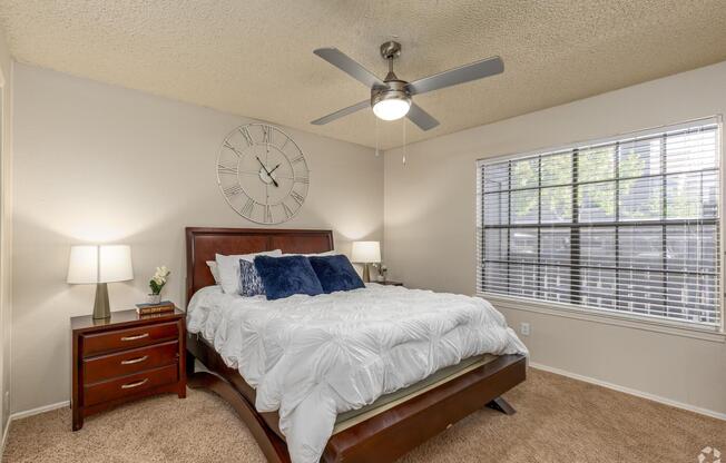A cozy bedroom featuring a large bed with white bedding and blue accent pillows. There are two bedside tables with lamps, a clock on the wall, and a window with blinds allowing natural light to enter. A ceiling fan is seen above, adding to the comfortable ambiance.