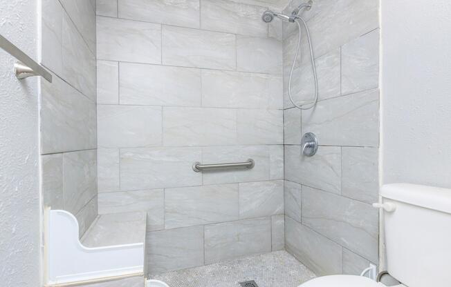A modern bathroom shower area featuring light gray tiled walls, a handheld showerhead, grab bar for safety, and a built-in shower bench. The floor is tiled with small, round stones, and there is a white toilet adjacent to the shower. The overall design emphasizes a clean and contemporary look.