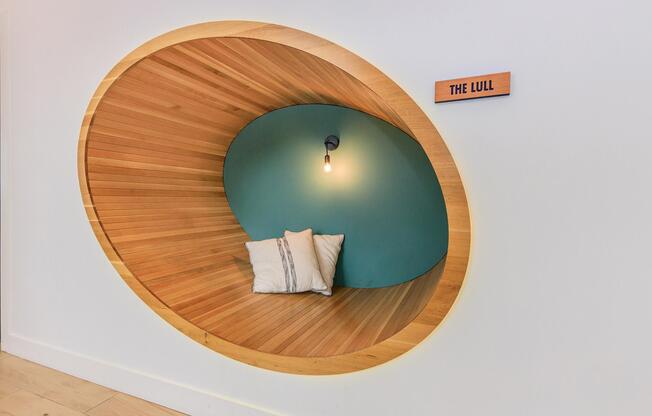 A cozy, circular nook in a wall featuring wooden slats and a teal backdrop. Inside the nook, there are two soft pillows, and overhead, a warm lightbulb hangs down, creating an inviting atmosphere. A wooden sign labeled "THE LULL" is mounted nearby, adding a touch of charm to the space.
