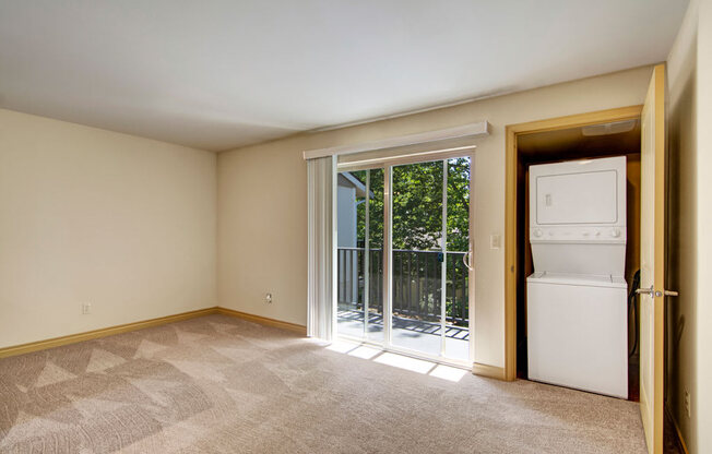 A spacious living room here at Asbury Park featuring soft neutral walls, plush carpet flooring, and a sliding glass door that opens to a private balcony with railing and leafy outdoor views, while a dedicated in-home laundry closet with stacked appliances is conveniently tucked behind a nearby door, offering a functional open layout with ample natural light.