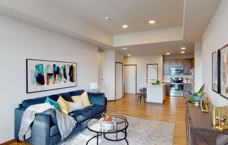 A stylish living room here at Van Buren East with wood-style flooring and modern furnishings. A cozy sofa and open layout flow into a kitchen with stainless steel appliances and a center island, creating a bright, inviting space.