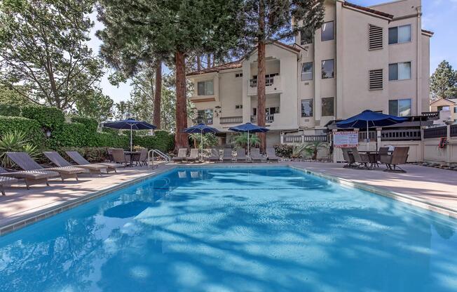 A serene swimming pool surrounded by lounge chairs and umbrellas, set against a backdrop of tall trees and a beige building. The clear blue water reflects the sunlight, creating a relaxing atmosphere perfect for leisure and enjoyment.