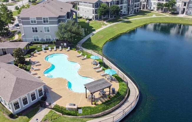A large swimming pool with a small waterfall and a hot tub is surrounded by a grassy area and a paved walkway.