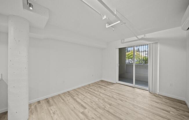 A bright, empty interior room with light wooden flooring, white walls, and a concrete column. There are large sliding glass doors leading to a balcony, allowing natural light to fill the space. The overall ambiance is modern and minimalist.