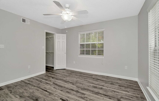 A well-lit, empty room with gray walls and wooden flooring. It features a ceiling fan, a window with blinds, and a closet with sliding doors. The space has a clean and modern aesthetic, making it suitable for various uses.