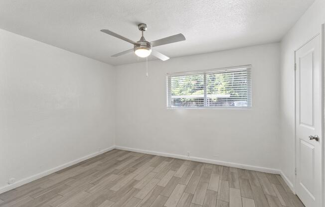 A bright, empty room with light-colored walls and tiled flooring. A ceiling fan is mounted in the center, and a window with blinds allows natural light to enter. The space is minimalistic, featuring no furniture, creating an open and airy feel.
