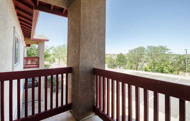A balcony with a red railing and a view of a parking lot and trees.