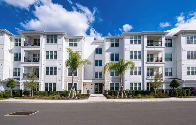 A row of white apartment buildings with balconies and trees in front.