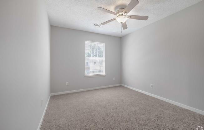 Empty room with light gray walls and carpet. A ceiling fan with five blades is mounted in the center, and there is a window on one wall allowing natural light to enter. The space is uncluttered and ready for furniture placement.