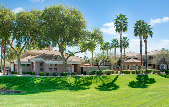 A scenic exterior view here at Dobson Towne Center showcasing lush green lawn, mature shade trees, and tall palm trees surrounding warm-toned residential buildings with tile roofs, complemented by neatly trimmed hedges and a peaceful, well-maintained landscape that creates an inviting and relaxed desert-inspired setting for residents.