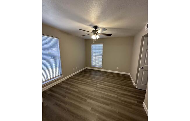 A small, minimally furnished room with light-colored walls and hardwood flooring. The room features a ceiling fan, two windows with blinds allowing natural light, and a door on the right side. The space is tidy and well-lit, suitable for various uses such as a bedroom or office.