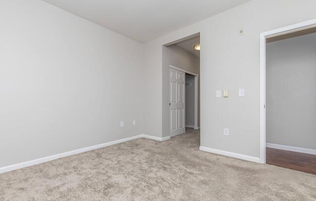 A sparsely furnished room with beige carpet and light gray walls. Includes a doorway leading to an adjacent space, a small closet, and two electrical outlets on the wall. Natural light from a ceiling fixture illuminates the area, creating a simple, clean ambiance.