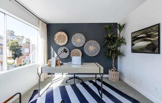 A modern office space featuring a sleek desk with a laptop and decorative items. The room has a large window with natural light, a striped rug on the floor, and a potted plant in the corner. Decorative wall hangings add a stylish touch to the gray accent wall. A framed picture is displayed on the wall.
