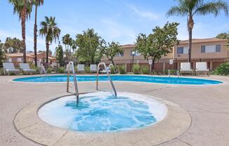 A serene outdoor swimming pool area featuring a large main pool and a smaller spa pool. Surrounding palm trees and well-maintained landscaping create a tropical atmosphere. Comfortable lounge chairs are arranged nearby, with residential buildings in the background under a clear blue sky.