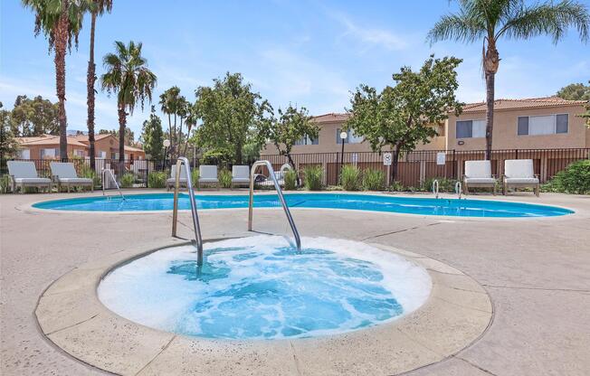 A serene outdoor swimming pool area featuring a large main pool and a smaller spa pool. Surrounding palm trees and well-maintained landscaping create a tropical atmosphere. Comfortable lounge chairs are arranged nearby, with residential buildings in the background under a clear blue sky.